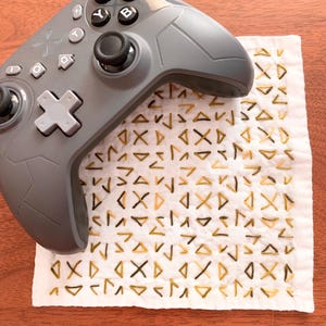 以下が含まれることがあります： グレーのゲームコントローラーが、金と茶色の幾何学模様の白い布の上に置かれています。 コントローラーには黒いボタンとグレーのアクセントがあります。 布は正方形で、手作りのようです。