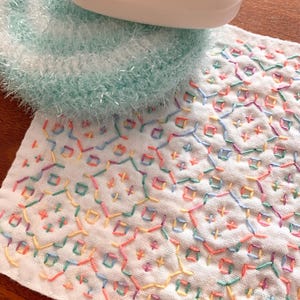 以下が含まれることがあります： 白い石鹸が、水色のふわふわのスクラビーと、カラフルな刺繍の幾何学模様が施された白い布の上に置かれています。布にはピンク、黄色、緑、青の色合いのステッチが施され、テクスチャードデザインを作り出しています。