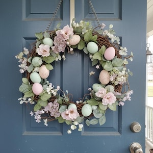 Peut inclure: Une couronne de Pâques décorative avec une base en vigne, ornée d'œufs aux couleurs pastel, de fleurs roses et blanches et de feuillage vert. La couronne est suspendue à une porte bleue, créant une ambiance printanière.