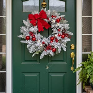 Op de afbeelding: Een feestelijke kerstkrans met bevroren witte takken, rode ornamenten, zilveren ballen en dennenappels. Een grote rode strik staat bovenaan. De krans hangt aan een donkergroene deur.