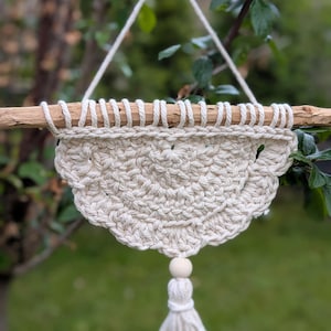 Könnte beinhalten: Cremefarbener Makramee-Wandbehang mit einem Holzdübel und einer Quaste. Das handgefertigte Stück hat ein strukturiertes, wellenförmiges Design und eine Holzperle. Die Wanddekoration wird von einer weißen Schnur gehalten, ideal für Boho-Stil.