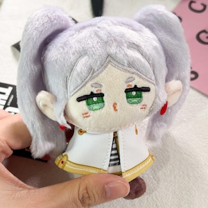 May include: A plush doll featuring light lavender hair styled in two side ponytails, embroidered green eyes, and pointed ears. The doll wears a white jacket with gold trim and a black and white striped shirt underneath, accented by small red circular earrings.