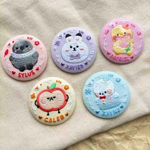 May include: Five embroidered circular patches with cartoon animal designs. The patches feature a gray owl, a white bunny, a yellow chick, a red apple, and a white bear. Each patch has a name embroidered on it: Sylus, Xavier, Rafayel, Caleb, and Zayne.