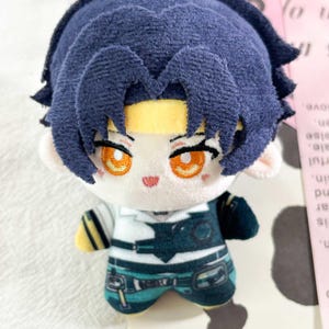 May include: A plush doll featuring dark blue hair, a yellow headband, and bright orange embroidered eyes. The doll wears a white shirt with a dark teal vest, black straps, and teal pants with a printed belt design. The plush has a soft texture.