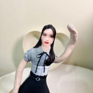 以下が含まれることがあります： 長い黒髪の人物がプリントされたカスタム布製人形。水色のピンストライプの半袖ブラウスに黒いリボン、ベルト付きの黒いスカートを着用している。人形は明るい色の腕を外側に伸ばしたポーズをとっている。