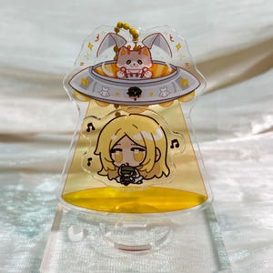 May include: A clear acrylic standee featuring a cartoon cat inside a silver UFO, with a yellow light beam shining down on a blonde character with musical notes floating nearby. The design includes small star accents and a gold ball chain attachment.