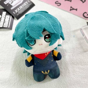Peut inclure: Une poupée en peluche avec des cheveux bleu sarcelle, des yeux brodés aux pupilles rouges et une petite bouche. La poupée porte une tenue bleu foncé avec des accents jaunes et un col rouge. Le tissu est doux et le design inclut un petit emblème doré sur la poitrine du costume bleu foncé.