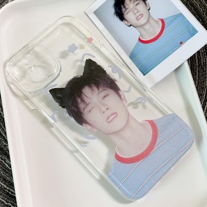 May include: A clear phone case featuring a printed graphic of a person with black cat ears, wearing a light blue and white striped shirt with a red collar. The design includes small light blue and pink star and swirl accents on the transparent surface.