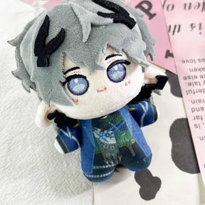 May include: A plush doll with short gray hair, black hair accents, and embroidered blue eyes. The doll features pointed ears, small black wings, and wears a blue and green patterned outfit with intricate details. The doll has a soft texture.