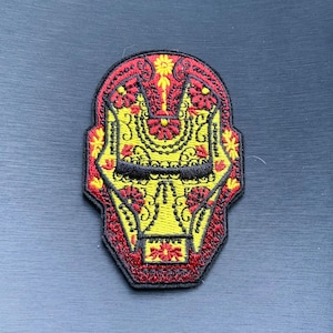 May include: Embroidered patch featuring a stylized Iron Man mask design. The mask is primarily yellow with red and black accents, incorporating floral patterns. The patch has a black border.