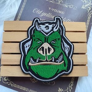 May include: Embroidered patch depicting a green orc face with tusks, a beige snout, and white details. The patch has a black border and is set against a wooden background. The text "Y-YOUMEI" is visible.