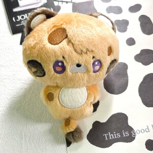 May include: A plush toy featuring a tan, fuzzy body with dark brown spots, a white circular belly patch, and purple embroidered eyes. The character has small rounded ears, a light gray muzzle, and a small tail with a dark brown tip.