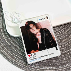 May include: A rectangular acrylic keychain featuring a social media post design with a portrait of a person in a black top. The graphic includes a profile name, a heart icon with 0,411 Likes, and Chinese text. A white star-shaped attachment is included.