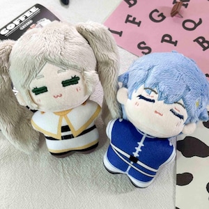 May include: Two plush dolls with embroidered facial features. The left doll has light beige hair in pigtails and wears a white outfit with yellow and black accents. The right doll has light blue hair and wears a blue outfit with white stripes.
