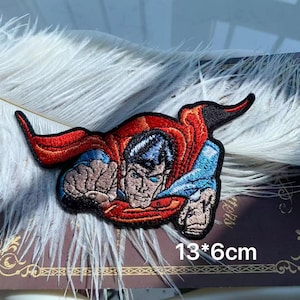 Könnte beinhalten: Gestickter Superman-Aufnäher, der den Superhelden in einer dynamischen Pose zeigt. Der Aufnäher zeigt Supermans klassischen roten Umhang, blauen Anzug und entschlossenen Gesichtsausdruck. Größe: 13 cm x 6 cm.