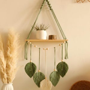 May include: A macramé wall shelf with a wooden shelf and hanging green leaves. The shelf is suspended by green macramé rope and features a small white pot with a plant and a small wooden block. Decorative and bohemian.