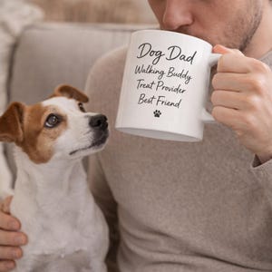 Puede incluir: Una taza de cerámica blanca con las palabras "Dog Dad, Walking Buddy, Treat Provider, Best Friend" impresas en negro, junto con una huella de pata. Un perro Jack Russell Terrier mira la taza.