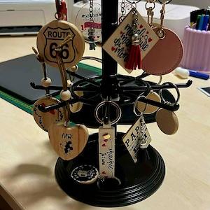 May include: A black metal rotating display stand holding various keychains. The keychains are made of wood and leather, with designs including Route 66, hearts, and text. Some keychains have gold key rings.