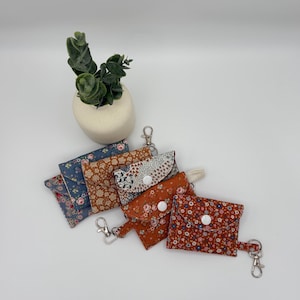 Könnte beinhalten: Eine Sammlung von fünf kleinen Stoffbeuteln mit Blumenmustern in Blau-, Orange- und Weißtönen. Jeder rechteckige Beutel verfügt über eine Klappe mit weißem Druckknopf und einen silbernen Karabinerhaken an einer seitlichen Lasche.