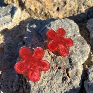 Puede incluir: Dos pendientes con forma de flor fabricados en resina roja translúcida y brillante. Cada pendiente presenta cinco pétalos redondeados con una textura sutil, brillante y arremolinada incrustada en el material rojo intenso, creando una superficie reflectante.