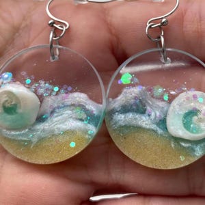 Puede incluir: Pendientes redondos y transparentes con temática de playa. Cada pendiente presenta una concha, arena y olas del océano con detalles de purpurina. Los pendientes tienen ganchos plateados.