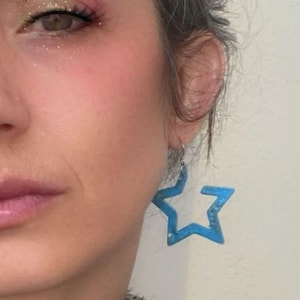 Puede incluir: Primer plano del rostro de una persona, con pendientes azules en forma de estrella. Los pendientes son de color azul claro. La persona lleva un collar de gargantilla negro y una chaqueta gris.