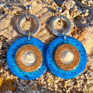 Puede incluir: Un par de pendientes colgantes con un anillo de metal plateado en la parte superior, conectado a un colgante circular más grande. El colgante consta de una base de resina azul brillante con purpurina, un anillo texturizado color arena y un centro de concha blanca.