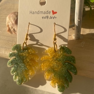 Puede incluir: Pendientes con forma de hoja con ganchos dorados. Los pendientes presentan un degradado de verde y amarillo, con forma de hojas de monstera. La tarjeta dice "Handmade with love".
