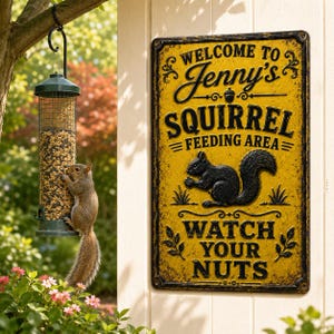 May include: A rectangular yellow metal sign with black text reading Welcome to Jenny's Squirrel Feeding Area Watch Your Nuts, featuring a black squirrel illustration. Beside it hangs a cylindrical bird feeder filled with mixed seeds.