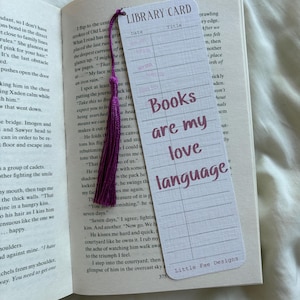 May include: A bookmark with a purple tassel is in an open book. The bookmark has a library card design with the words "Books are my love language" in a handwritten font. The bookmark is white with purple text. The book is open.
