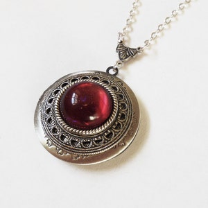 May include: A silver locket necklace with a red glass cabochon set in an ornate silver frame. The chain is silver and has a decorative clasp.