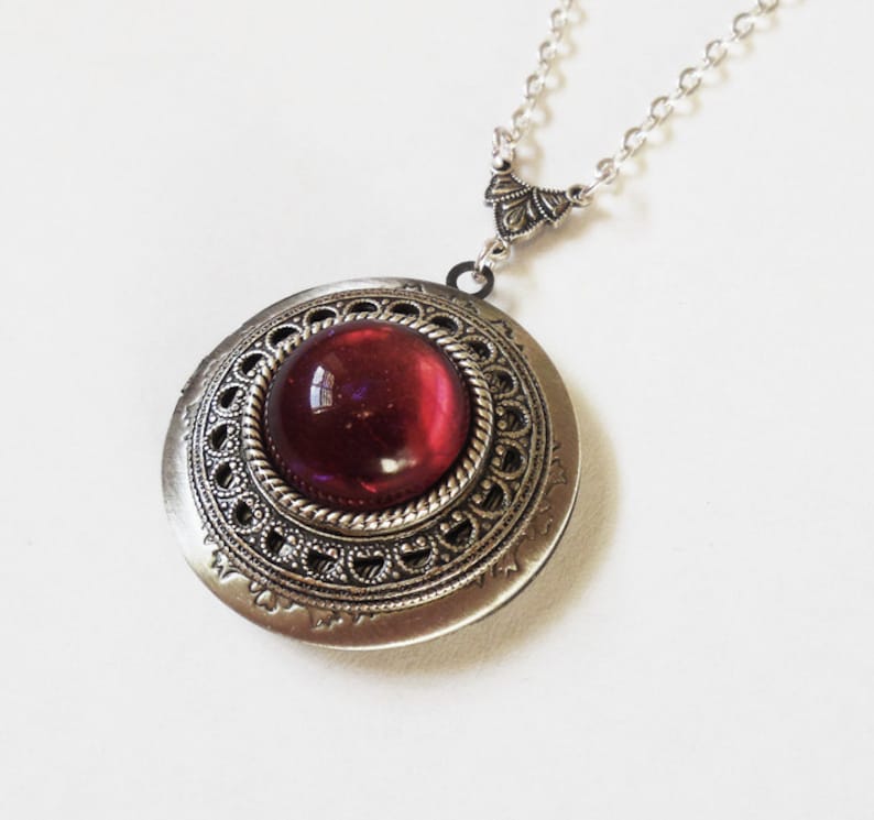 May include: A silver locket necklace with a red glass cabochon set in an ornate silver frame. The chain is silver and has a decorative clasp.