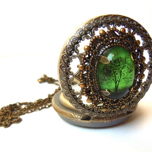 May include: An antique-style locket with a bronze-colored filigree design. The locket features an oval-shaped green glass insert with a tree silhouette. A bronze chain is attached to the locket.