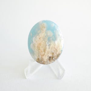 Pode incluir: Uma pedra preciosa oval com um padrão azul celeste e creme, que lembra nuvens. A pedra é exibida em um suporte de plástico transparente, sobre um fundo branco. O design da pedra preciosa evoca uma estética natural e arejada.