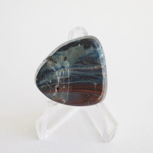 Könnte beinhalten: Ein polierter, abstrakter Stein mit einer Mischung aus blauen, braunen und weißen Farben. Der Stein wird auf einem transparenten Acrylständer präsentiert, der seine einzigartige Form und marmorierte Optik zeigt. Die Oberfläche des Steins hat ein glänzendes Finish.