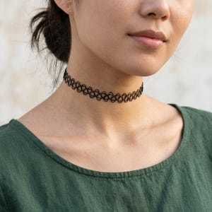 May include: A black tattoo-style choker necklace featuring an intricate, repeating woven pattern. The accessory is designed to fit snugly around the neck, showcasing a delicate, open-work structure that creates a distinct, textured appearance.