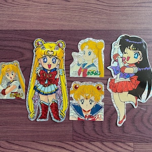 Puede incluir: Colección de cinco pegatinas brillantes de personajes de Sailor Moon. Las pegatinas presentan varios personajes con colores vibrantes, incluyendo amarillo, rojo, azul y blanco. Las pegatinas están sobre una superficie oscura con vetas de madera.