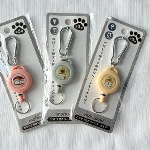 May include: Three retractable key holders in pink, gray, and light yellow, each featuring a mofusand cat illustration. The holders include a silver carabiner, a circular plastic housing, and a metal ring, all packaged on individual display cards.