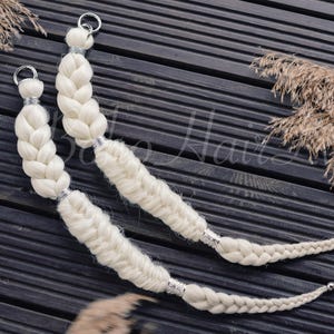 Puede incluir: Dos extensiones de cabello trenzado blanco roto con detalles plateados y anillos metálicos. Se encuentran sobre una superficie de madera oscura, con plantas secas en el fondo. Las trenzas son gruesas y texturizadas.