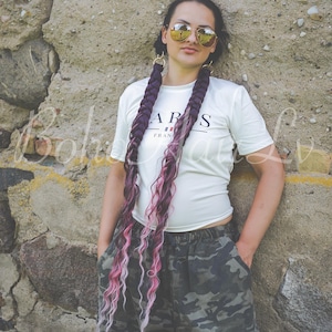Op de afbeelding: Een wit T-shirt met korte mouwen en de tekst PARIS FRANCE op de voorkant, gecombineerd met een broek met camouflageprint. De outfit bevat lange, dikke gevlochten haarextensies in donkerpaarse en roze tinten en een goudkleurige zonnebril.