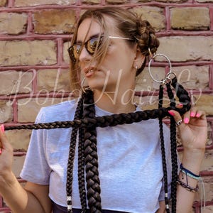 Puede incluir: Una camiseta gris claro combinada con extensiones de cabello sintético de color marrón oscuro peinadas en trenzas gruesas. Las trenzas están unidas a grandes anillos de metal plateado y tienen pequeñas cuentas cilíndricas plateadas en los extremos.
