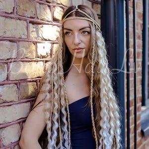 May include: A gold-toned metal headpiece featuring a central ring and multiple thin braided strands that drape down. The accessory is worn over long, wavy, blonde-highlighted hair, with the metallic braids integrated into the hair strands.
