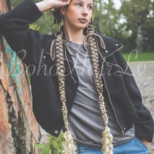 Op de afbeelding: Twee lange, dikke haarextensies met een gevlochten ontwerp dat verloopt van een lichtbruine tint aan de bovenkant naar een heldere blonde kleur aan de uiteinden, afgewerkt met losse, krullende lokken. De extensies zijn bevestigd aan goudkleurige ringen.