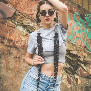 Puede incluir: Dos extensiones de cabello trenzado negro con anillos de metal plateado y cordones blancos. Las trenzas están unidas a grandes aros plateados diseñados para llevarse alrededor del cuello, cayendo sobre el pecho sobre un top corto gris.