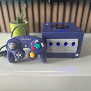 May include: A purple Nintendo GameCube console and controller. The console is cube-shaped with silver accents and three lights. The controller has a D-pad, analog stick, and coloured buttons. The text "NINTENDO GAMECUBE" is visible on the controller.