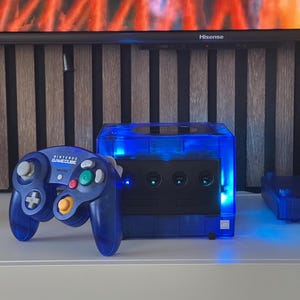 Puede incluir: Una consola Nintendo GameCube azul y un mando a juego. La consola tiene una carcasa azul transparente con un panel frontal negro. El mando también es azul con botones coloridos. La consola está sobre una superficie blanca.