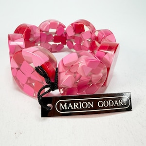 May include: A chunky bracelet composed of circular resin links filled with a mosaic of pink and red geometric fragments. A black string attaches a rectangular black tag with white text that reads MARION GODART to the bracelet.