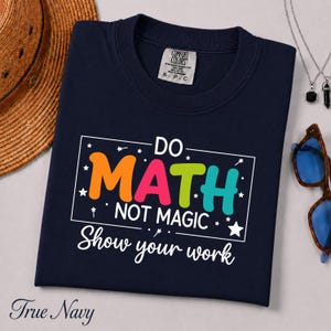 May include: A navy blue t-shirt featuring a white rectangular border containing the colourful text DO MATH NOT MAGIC in orange, pink, lime green, and teal, with the white text Show your work below and small star accents throughout the design.