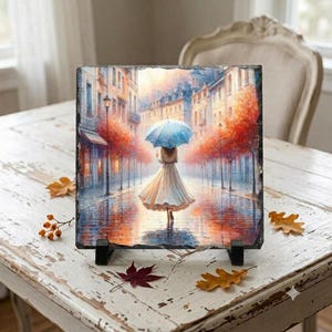 May include: A square slate plaque featuring a printed illustration of a person in a light-coloured dress holding a blue umbrella, walking down a city street lined with buildings and trees with orange and red autumn foliage during a rain shower.
