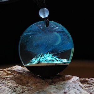 Può includere: Una collana con pendente rotondo raffigurante una scena di montagna in miniatura sotto un cielo stellato. Il pendente è realizzato in resina trasparente con un design blu e bianco e una base nera. La collana è appesa a un cordino nero.